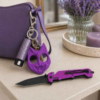 A purple folding survival knife with a black blade is displayed on a beige countertop next to a matching purple zippered purse, a purple cat-defense keychain, and a glittery purple pepper spray keychain, with a vase of lavender in the background.