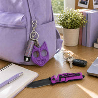A purple cat-shaped keychain and bling pepper spray with matching purple backpack are shown on a wooden desk beside a notebook, pen, folding knife, keys, and a potted plant.