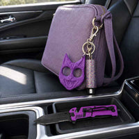 A purple textured leather crossbody bag with a matching purple cat-defense pepper spray keychain and a silver key fob, shown in a car's center console next to a purple folding knife.
